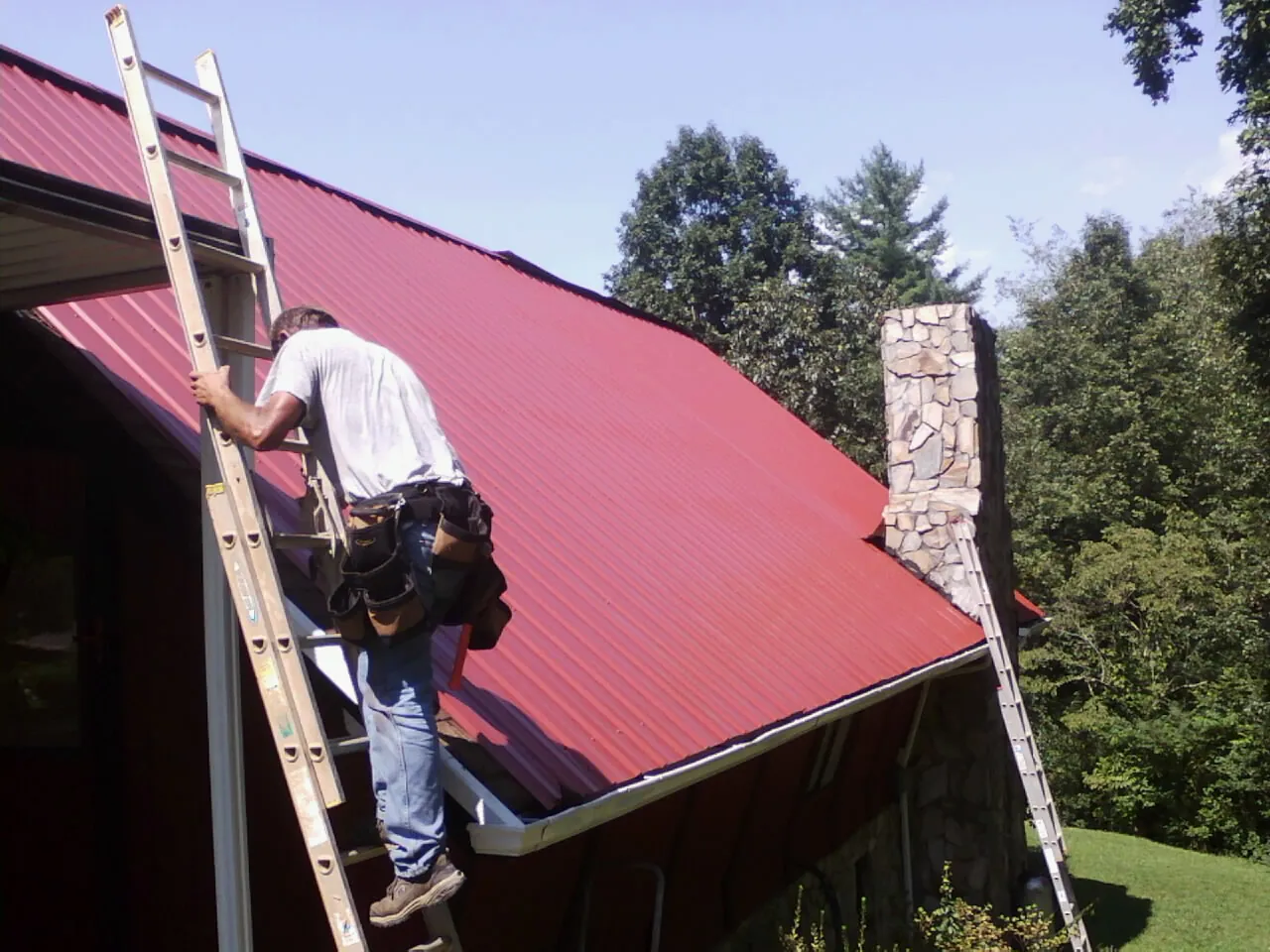 Roofer on a ladder beside a red metal roof and stone chimney — Soffit & Fascia Repair by Roofing Brothers in Ash